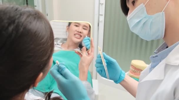 アジアの男性歯科医は 歯科医院 歯科衛生 病院の専門医療医でインプラント歯科色合いの若い女性患者に矯正ケアとホワイトニングを説明しています — ストック動画