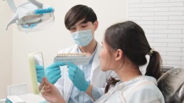 Asyalı erkek diş hekimi diş sağlığı kliniğinde takma diş tonu, diş hijyeni ve hastanede uzman sağlık uzmanı olan genç bir kadına diş bakımı ve beyazlatmayı açıklıyor..