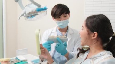 Asyalı erkek diş hekimi diş sağlığı kliniğinde takma diş tonu, diş hijyeni ve hastanede uzman sağlık uzmanı olan genç bir kadına diş bakımı ve beyazlatmayı açıklıyor..