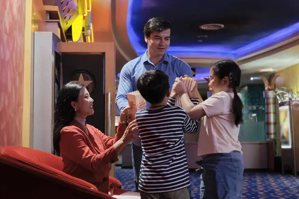 Cheerful Asian family at cinema show. Mum and kids sit in theater seat, waiting for father\'s popcorn before watching movie, and happy together, a public indoor entertainment lifestyle with film shows.
