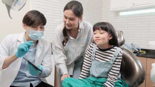 アジアの男性小児歯科医が歯科医院のタブレットからの歯科衛生のための少女および彼女の母に歯X線スキャン 子供の医療病院の専門の矯正口頭医を説明します — ストック動画