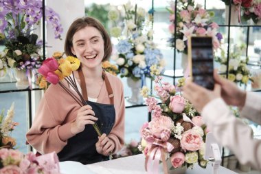 E-commerce business, one young White female florist demonstrates and shows floral arrangements via online live streaming with smartphone application in bright flower shop, beautiful blossoms store. 