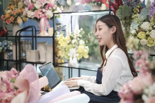 One young Asian female florist and SME entrepreneur in apron, works with laptop, online selling floral arrange in colorful flower shop store with beautiful bunch of blossoms, and e-commerce business.