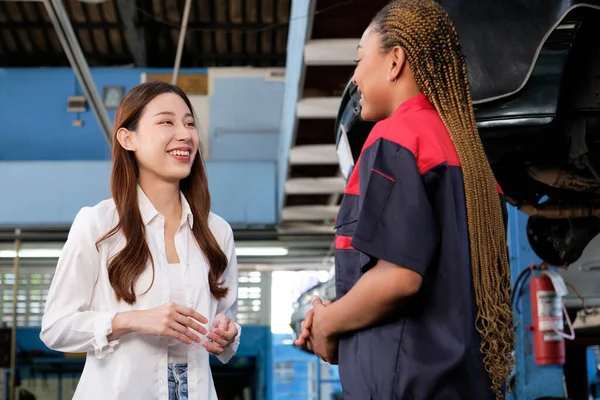 Asian female customer talking with Black automotive mechanic worker, quality guarantee with trust at maintenance garage, professional vehicle service station, ensuring fix checks, and repair industry.