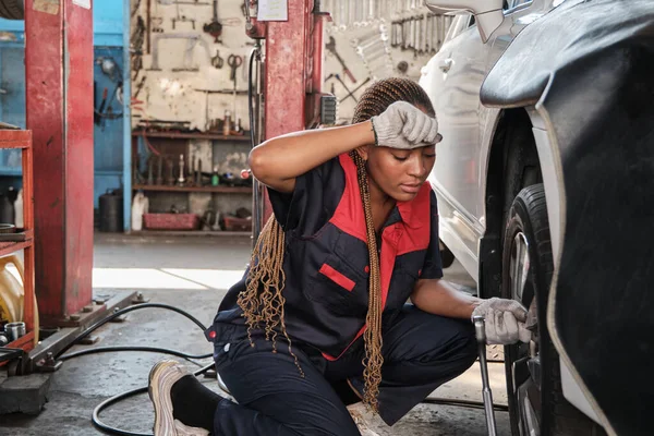 Black female automotive mechanic worker screws car wheel nuts with wrench for repair at garage, wipes off sweat, tired with labor works, vehicle maintenance service jobs, industry occupation business.