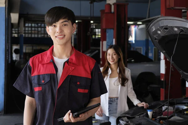 Portrait of young Asian male automotive mechanic worker with female customer\'s EV car at maintenance garage, happy smiles, professional service station, check and repair in auto transport industry.