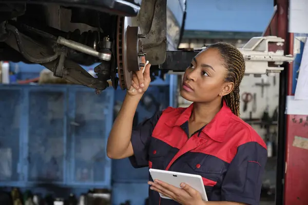 Expert young Black female automotive mechanic worker checks car\'s wheel brake disc and repairs inspect checklist by tablet at fix garage. Vehicle maintenance service works industry occupation jobs.