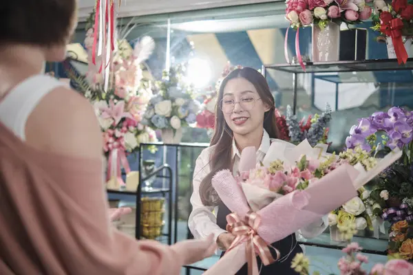 Young Asian female florist worker in apron delivers beautiful fresh blossoms bouquet to customer who purchased order, happy seller who works in colorful flower shop, and small business entrepreneur.