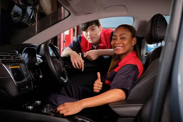 Young Black female automotive mechanic technician and partner check maintenance list in car interior at garage with smiles. Vehicle service fix and repair works, industrial occupation business jobs.