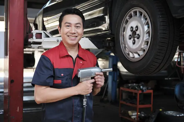 One expert Asian male automotive mechanic technician is screwing car wheel nuts on lifting with drill wrench for repair at garage. Vehicle maintenance service works industry occupation business jobs.