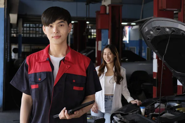 Portrait of young Asian male automotive mechanic worker with female customer\'s EV car at maintenance garage, happy smiles, professional service station, check and repair in auto transport industry.
