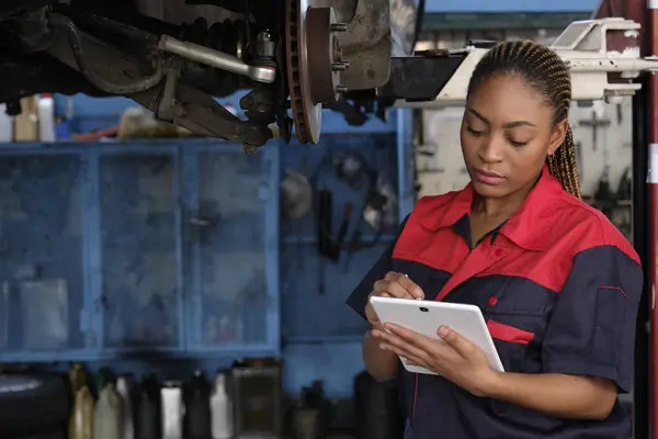 Expert young Black female automotive mechanic worker checks car's wheel brake disc and repairs inspect checklist by tablet at fix garage. Vehicle maintenance service works industry occupation jobs.