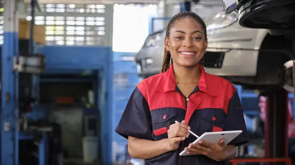 Happy Black female supervisor mechanic, cheerful smile, inspects repair work checklists with tablet at garage, service car maintenance, and fixing specialist occupations in auto transport industry.