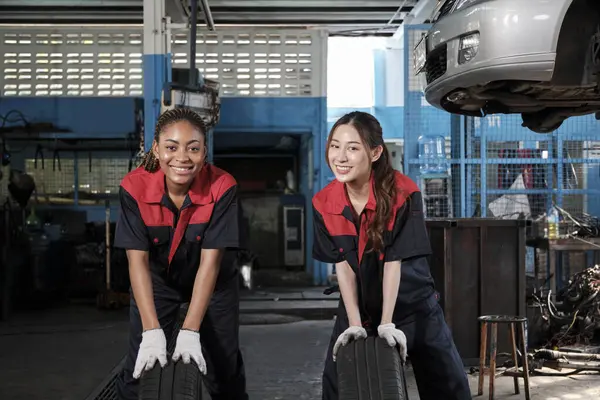 Two diverse female automotive mechanic worker partners compete to push car wheels for change and repair at garage, vehicle maintenance service jobs, industry occupation, and professional labor works.
