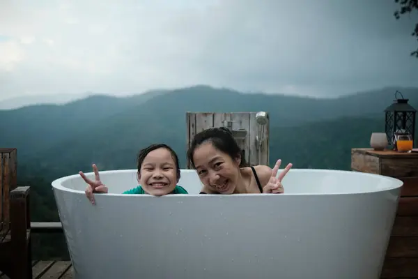 Asian family, mum and child playing together, leisurely relax in bathtub, enjoy happily with panoramic mountain range view of tropical landscape in natural scenery travel resort for holiday vacation.