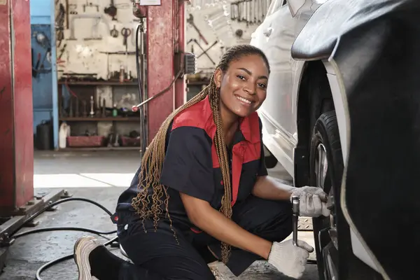 Black female automotive mechanic worker screws car wheel nuts with wrench for repair at garage, vehicle maintenance service jobs, industry occupation business, happy labor works, smile and cheerful.