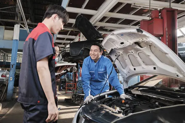 Asian male professional automotive engineer supervisor describes car engine maintenance and repair work with mechanic worker staffs team in fix service garage, specialist occupations in auto industry.