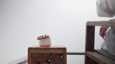 Yakın çekim, dişi turist eli ahşap masaya bir fincan sıcak çay koyuyor, sabah sıcak, serinlemek için yudumluyor ve doğal tatil beldesindeki sakin sis manzarasında dinleniyor, boş zamanlarında seyahat ediyor ve tatilde dinleniyor..