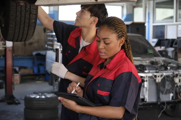 Young expert Black female inspects repair checklist with automotive mechanic worker partner, quality suspension technician team at fix garage. Vehicle maintenance service works industry occupation jobs.