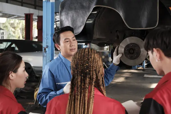 Asian male professional automotive engineer supervisor describes car wheel and suspension repair work with mechanic worker staffs team in fix service garage, specialist occupations in auto industry.