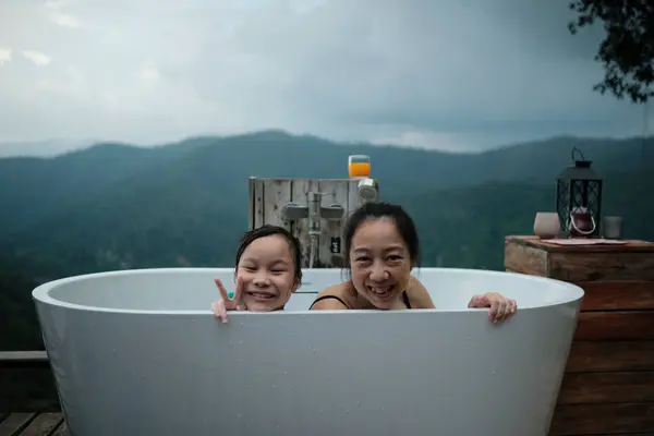 Asian family, mum and child playing together, leisurely relax in bathtub, enjoy happily with panoramic mountain range view of tropical landscape in natural scenery travel resort for holiday vacation.