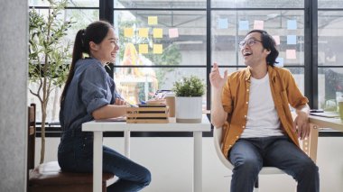 İki Asyalı işçi çalışma masalarında oturuyor, iş birliği yapıyor ve günlük iş ofislerinin pencerelerinde konuşuyorlar, online iş arkadaşları, pozitif ilişkiler.