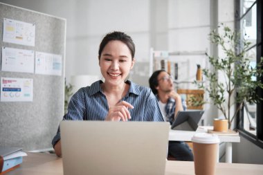 Güzel genç bayan Asyalı işçi çalışmaktan, dizüstü bilgisayar kullanmaktan, masaya gülümsemekten, fikirleri düşünmekten ve günlük ofis ve başlangıç KOBİ 'sinde yaratıcı pazarlama işlerinde yoğunlaşmaktan mutlu..