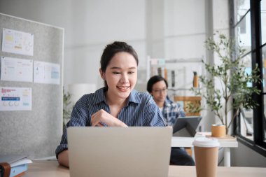 Güzel genç bayan Asyalı işçi çalışmaktan, dizüstü bilgisayar kullanmaktan, masaya gülümsemekten, fikirleri düşünmekten ve günlük ofis ve başlangıç KOBİ 'sinde yaratıcı pazarlama işlerinde yoğunlaşmaktan mutlu..