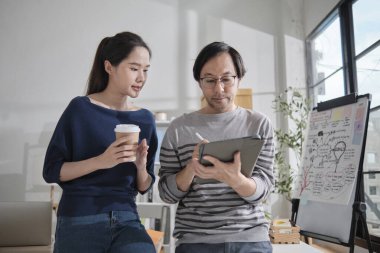 İki genç Asyalı girişim ortağı gelişigüzel küçük işletme ofisinde tartışıyor ve fikir alışverişinde bulunuyor, bir tablet üzerinde pazarlama planlama işleri ve E-ticaret girişimcisi ve iş arkadaşı olarak serbest çalışma.