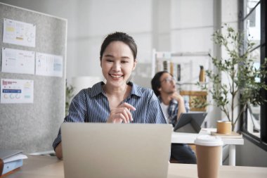 Güzel genç bayan Asyalı işçi çalışmaktan, dizüstü bilgisayar kullanmaktan, masaya gülümsemekten, fikirleri düşünmekten ve günlük ofis ve başlangıç KOBİ 'sinde yaratıcı pazarlama işlerinde yoğunlaşmaktan mutlu..