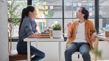 İki Asyalı işçi çalışma masalarında oturuyor, iş birliği yapıyor ve günlük iş ofislerinin pencerelerinde konuşuyorlar, online iş arkadaşları, pozitif ilişkiler.