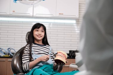 Asyalı kız dişlerini pediatrik bir dişçiye fırçaladı, diş fırçası ve takma dişlerle diş kliniğinde ve çocuk hastanesinde ağız hijyenik ortodontik sağlık hizmetlerinde gösterdi..