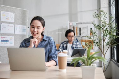 Güzel genç bayan Asyalı işçi çalışmaktan, dizüstü bilgisayar kullanmaktan, masaya gülümsemekten, fikirleri düşünmekten ve günlük ofis ve başlangıç KOBİ 'sinde yaratıcı pazarlama işlerinde yoğunlaşmaktan mutlu..