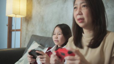 Asyalı aile, anne ve çocuk birlikte kanepede neşe kontrolörleriyle VDO oyunu oynuyorlar. Keyifli ve neşeli hafta sonu çocuk ve ebeveynlerin heyecanlı hobisi dijital eğlence..