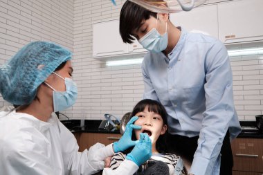 Asyalı kadın pediatrik diş hekimi bir kızın dişlerini diş kliniğinde muayene eder ve bir çocuk hastanesinde babaya, ağız hijyenine ve profesyonel ortodontik sağlık hizmetlerine özen gösterir..