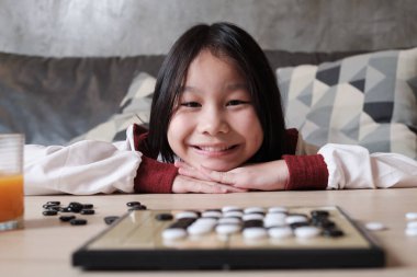 Portre Yakın plan, Go oynayan bir Asyalı kız, oturma odasındaki masada dama masası oyunu, neşeli gülümseme ve kameraya bakma, akıllı şirin çocuk kazananın rahatlatıcı hafta sonu hobisi.