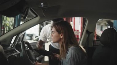 Siyah erkek satış müdürü ve satıcı, iç konsol teknolojisini otomotiv servis garajı, makine endüstrisi ve taşımacılık sektöründeki yeni EV arabadaki beyaz kadın alıcıya açıklıyor..