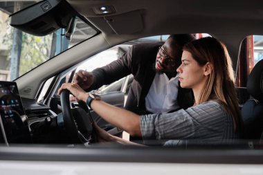 Siyah erkek satış müdürü ve satıcı, iç konsol teknolojisini otomotiv servis garajı, makine endüstrisi ve taşımacılık sektöründeki yeni EV arabadaki beyaz kadın alıcıya açıklıyor..