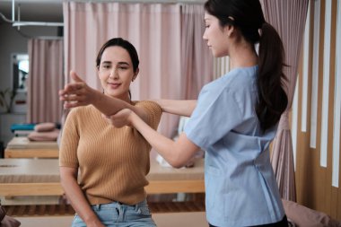 İspanyol kadın hasta kol esneme, tedavi, omuz kası ağrısı, Asyalı fizyoterapist tarafından tedavi, pasif tıbbi tedavi, sağlık masajı, spa kliniği.