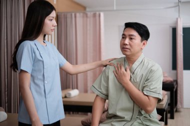 Olgun Asyalı hasta adam, pasif tıbbi tedavi sağlık merkezinin ortopedik koğuşunda omuz kası ağrısının iyileşmesi için kadın fizyoterapist doktoruna danışmanlık yapıyor..