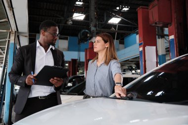 Garanti hizmeti işi, beyaz kadın müşteri araba boyasını inceliyor ve siyah erkek sigorta acentesiyle tırmalama onarımı yapıyor, araba taleplerini kabul ediyor, araba tamirhanesinde tamirat ve bakım yapıyor..