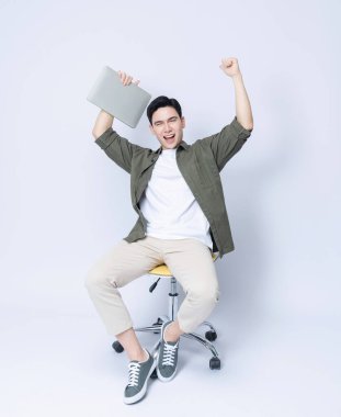 Genç Asyalı iş adamı sandalyede oturuyor ve arka planda dizüstü bilgisayar kullanıyor.