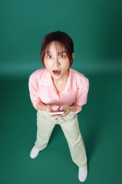 Yeşil arka planda akıllı telefon kullanan Asyalı genç kızın tam boy fotoğrafı.