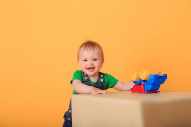 Küçük yakışıklı bir çocuk açık renkli bir puffe üzerinde bir oyuncakla oynuyor. Sarı arka planda bir çocuğun stüdyo çekimi..