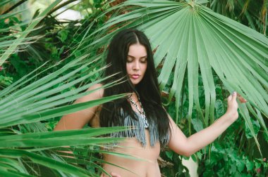 Palmiye yapraklarının yanında esmer bir kadının portresi. Bitkilerin arasında mayo giymiş, tropik iklimli bir kız.