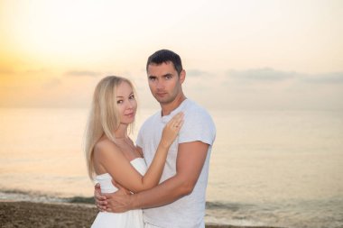 Deniz günbatımında ya da gün doğumunda arka planda kalan genç bir çift. Genç kadın ve genç esmer adam deniz kıyısında çakıl taşları üzerinde.