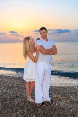 Deniz günbatımında ya da gün doğumunda arka planda kalan genç bir çift. Genç kadın ve genç esmer adam deniz kıyısında çakıl taşları üzerinde.
