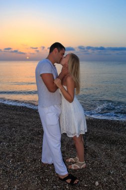 Deniz günbatımında ya da gün doğumunda arka planda kalan genç bir çift. Genç kadın ve genç esmer adam deniz kıyısında çakıl taşları üzerinde.