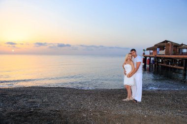 Deniz günbatımında ya da gün doğumunda arka planda kalan genç bir çift. Genç kadın ve genç esmer adam deniz kıyısında çakıl taşları üzerinde.