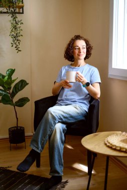 Pencerenin yanındaki koltukta oturan genç ve güzel bir kadın elinde bardak ya da kupayla. Yüksek kalite fotoğraf